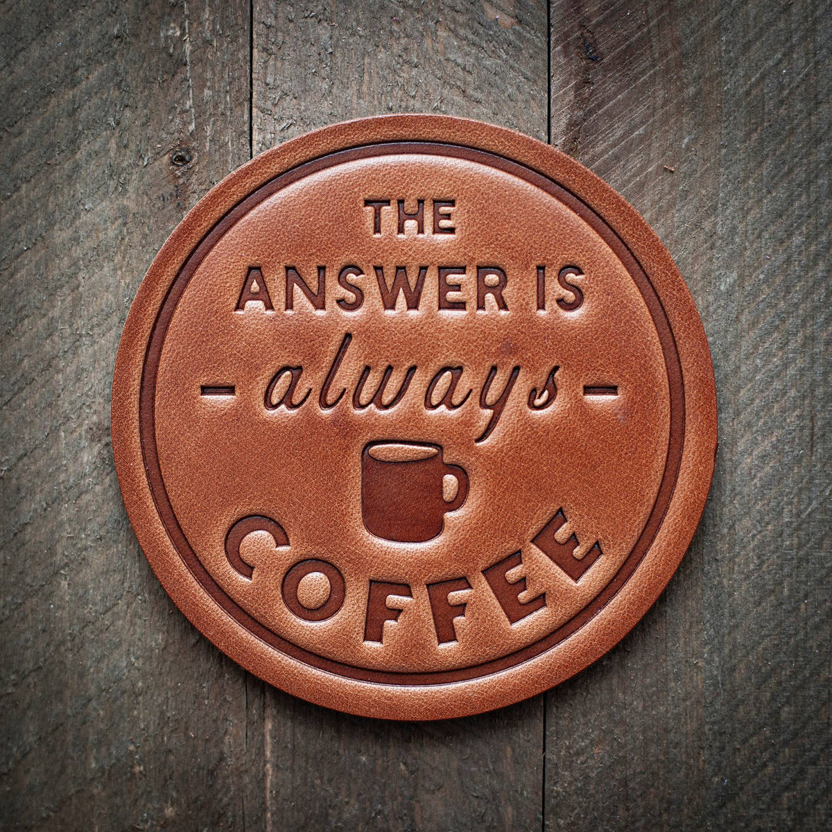 A brown leather coaster with the text 'THE ANSWER IS ALWAYS COFFEE' embossed on it, placed on a wooden surface.