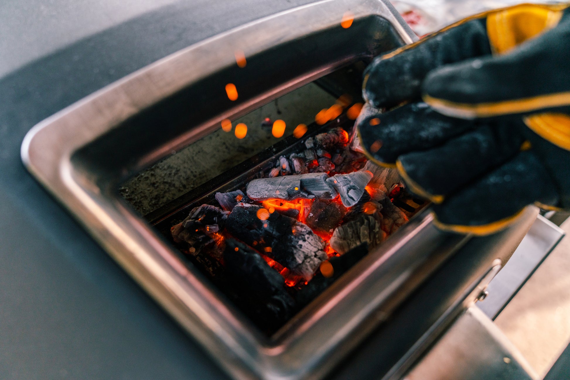 This close-up image shows wood that has burned down inside the oven. A person is wearing Ooni heavy gloves for safety while adding more wood.