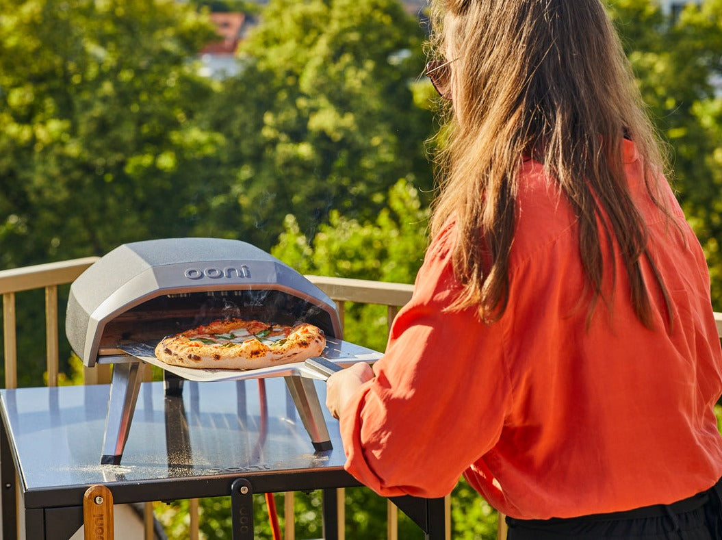 A woman smoothly pulls her perfect, spotted pizza from her Ooni oven.